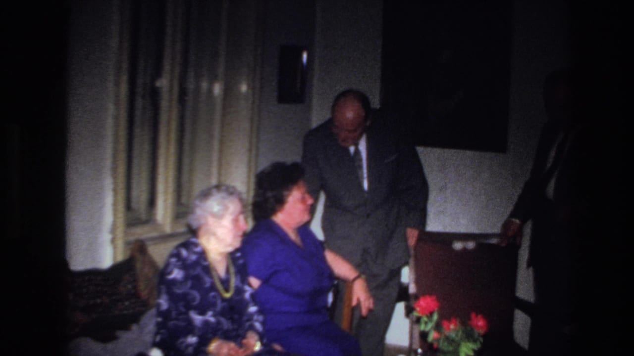 In 1967 France home-movie footage, two seated women look toward a suited man leaning forward near curtains and red flowers.