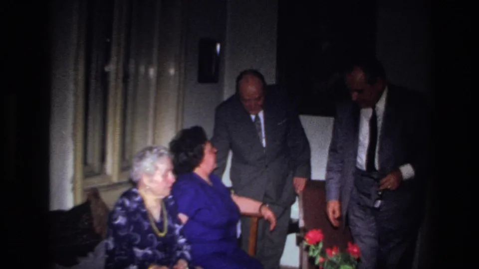 In the dim 1967 France home interior, a second suited man joins the seated women and standing man near a vase of red flowers.