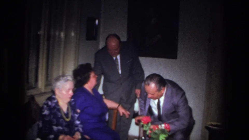 A suited man bends to examine or handle red flowers as two women sit and another man stands in the 1967 France interior.