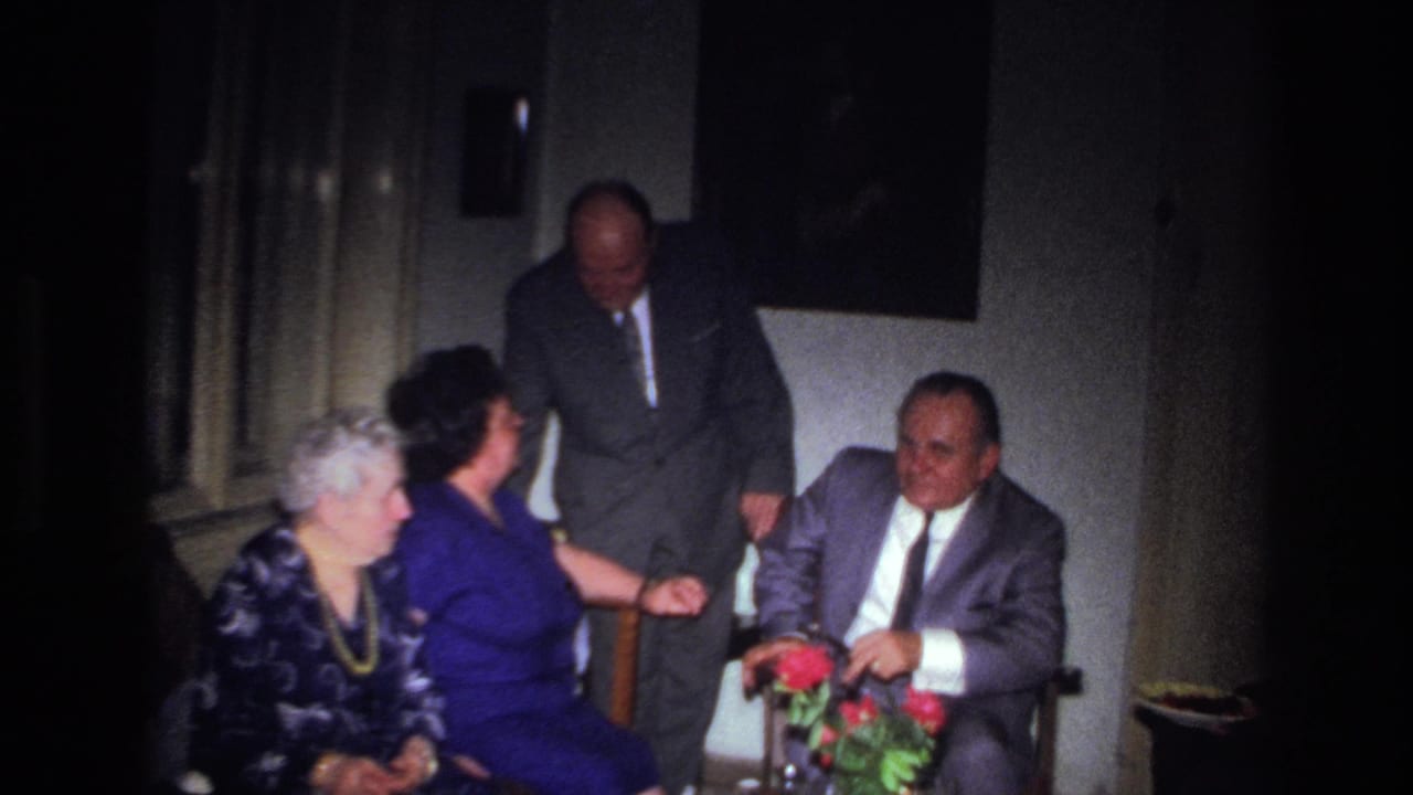1967 France home-movie frame of older adults in formal attire gathered indoors near a small table with red flowers.