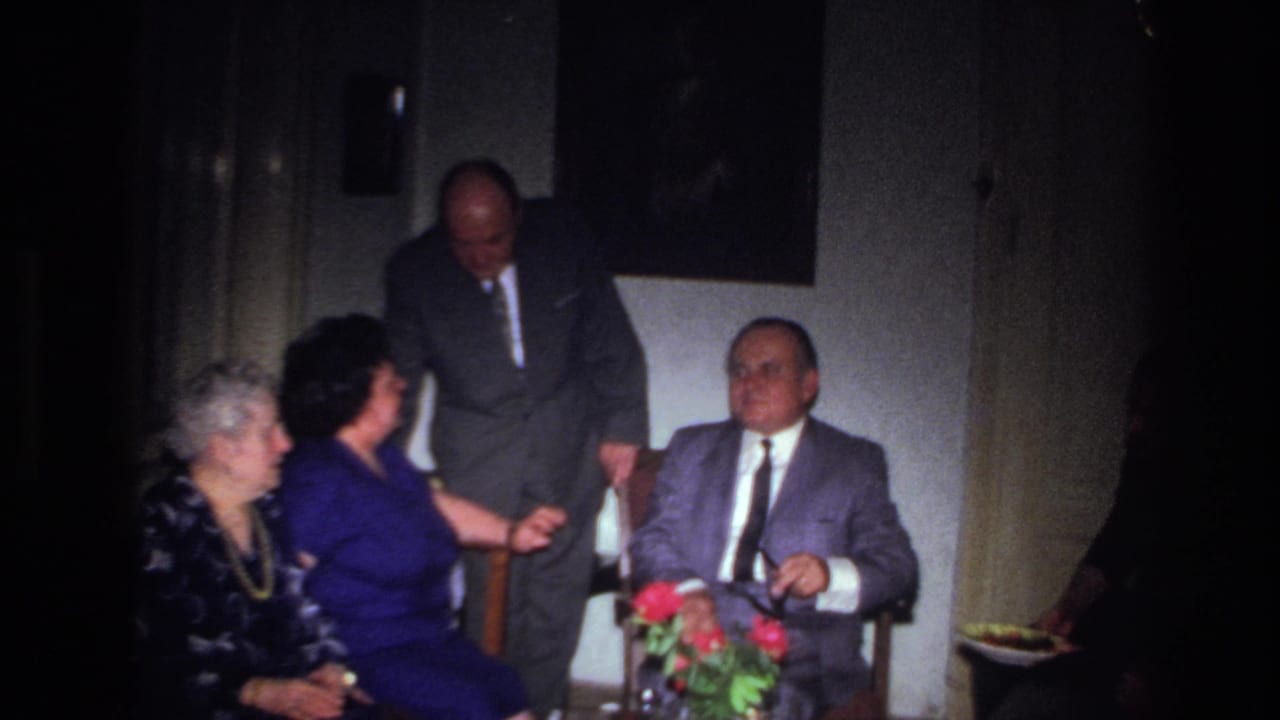 In a 1967 France interior, a standing man in a dark suit speaks near a seated man as two women sit beside flowers.