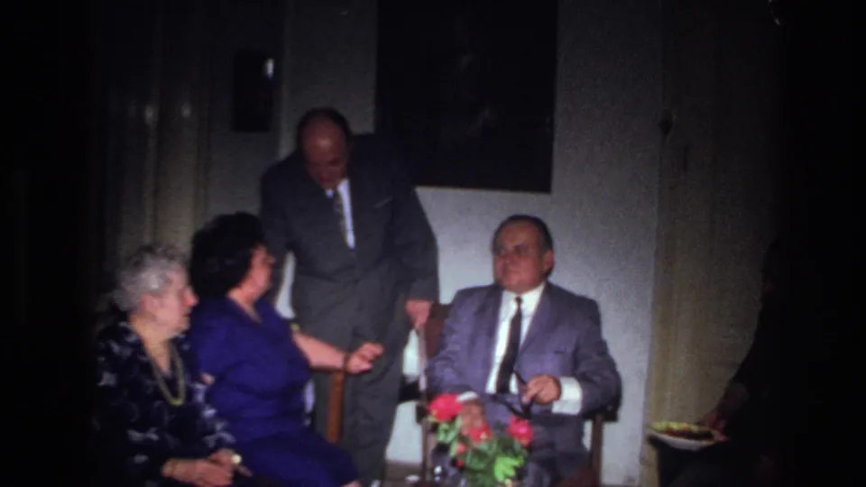 In a 1967 France interior, a standing man in a dark suit speaks near a seated man as two women sit beside flowers.