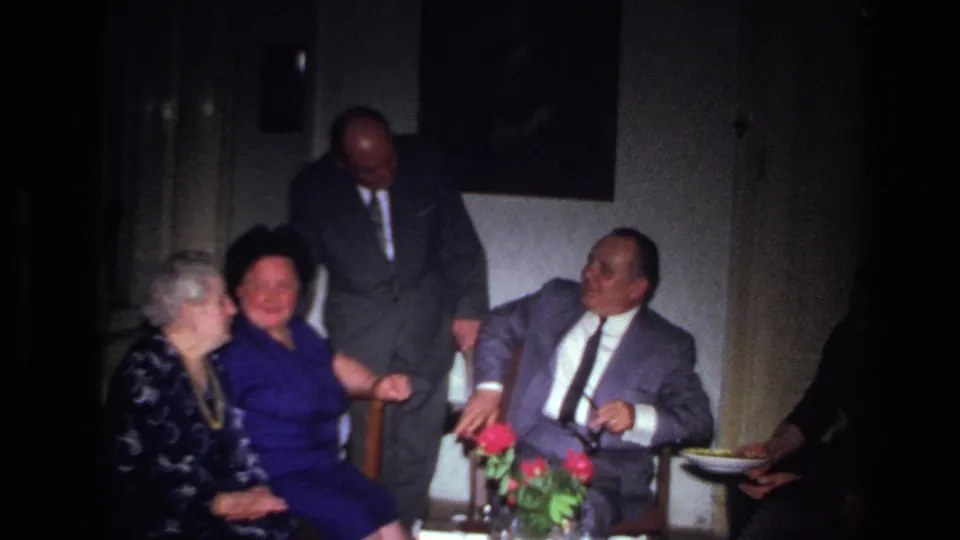 Older adults in formal dress are seen in a private French interior, with red flowers on a table during a solemn gathering.