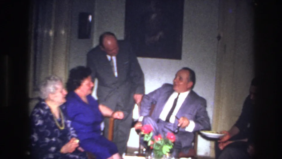 A 1967 France home-movie frame shows a plate offered at the edge of a solemn indoor gathering of older adults.