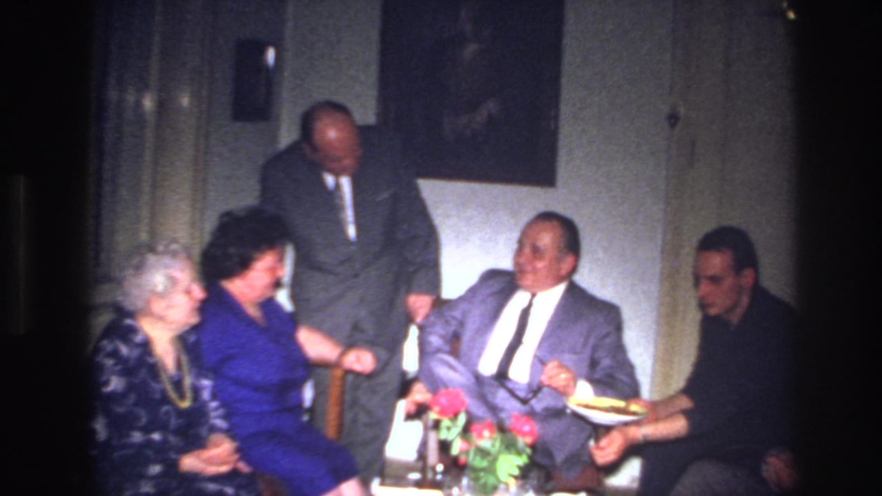 Five adults gathered in a dim living room as food is offered near a small table with red flowers.