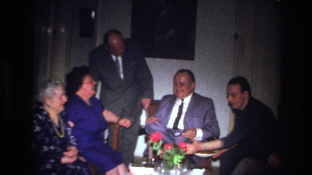Adults converse indoors while a seated man in a gray suit receives a plate from a man at right.