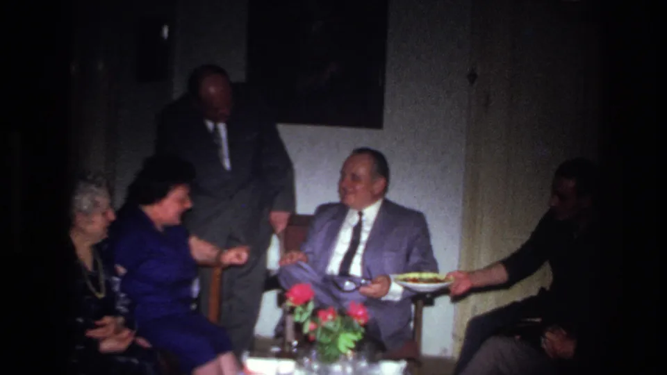 Soft 8mm frame from a 1967 French interior gathering, with adults seated near flowers and food being passed.