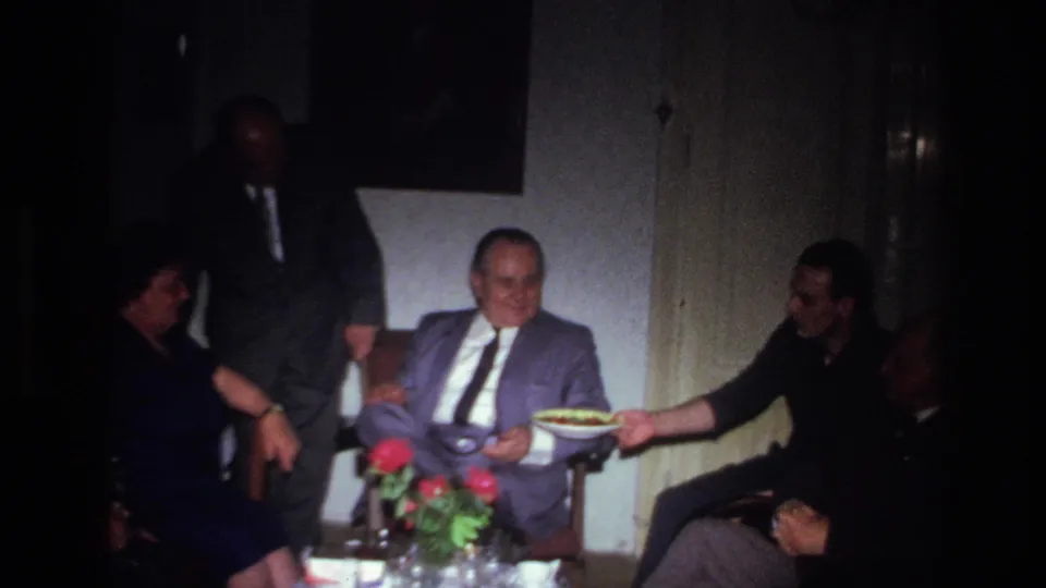1967 French home-movie frame of a seated older man smiling while food is offered during a private celebration.