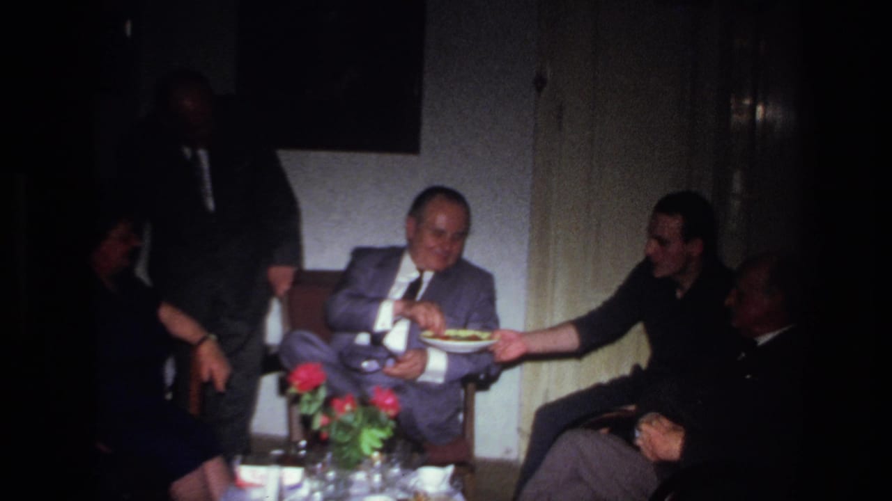 1967 France home-movie frame showing adult men seated indoors while a plate of food is passed toward a man in a gray suit.
