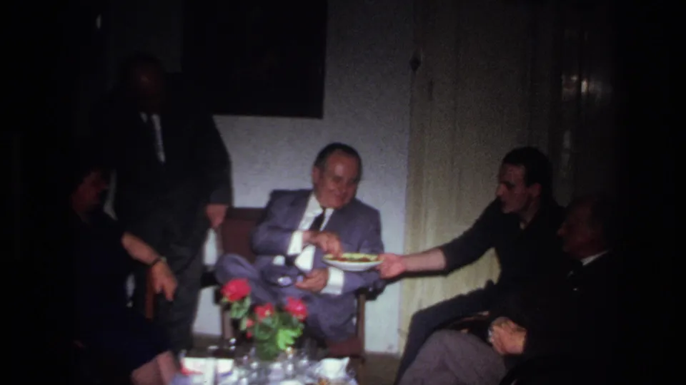 1967 France home-movie frame showing adult men seated indoors while a plate of food is passed toward a man in a gray suit.