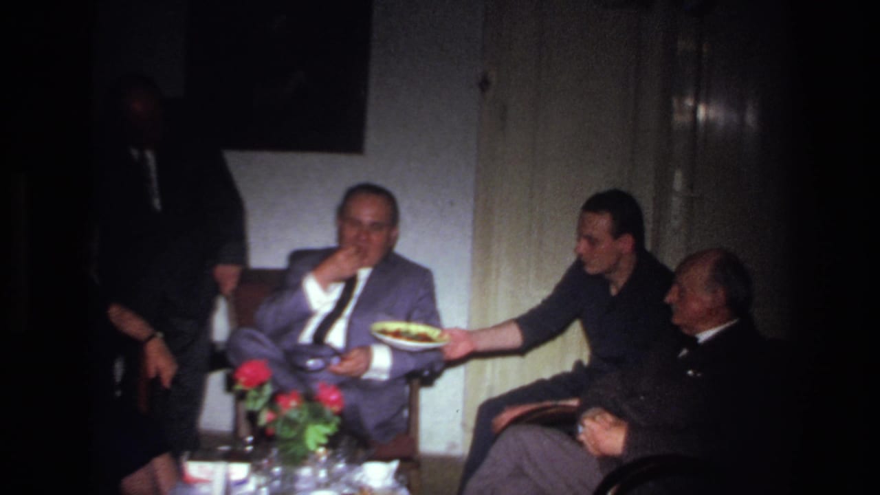 A man in a gray suit eats while another seated man holds a plate toward him.