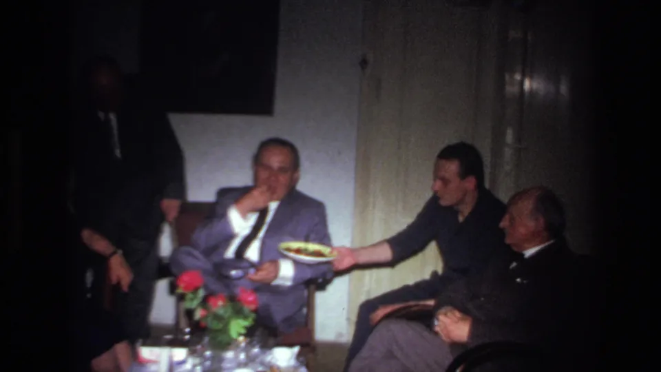1967 France home-movie frame of men seated close together indoors, with one man eating and another holding out a plate.