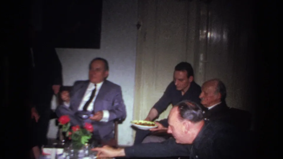 1967 France home-movie frame showing a seated group of adult men around a small table with red roses and food nearby.