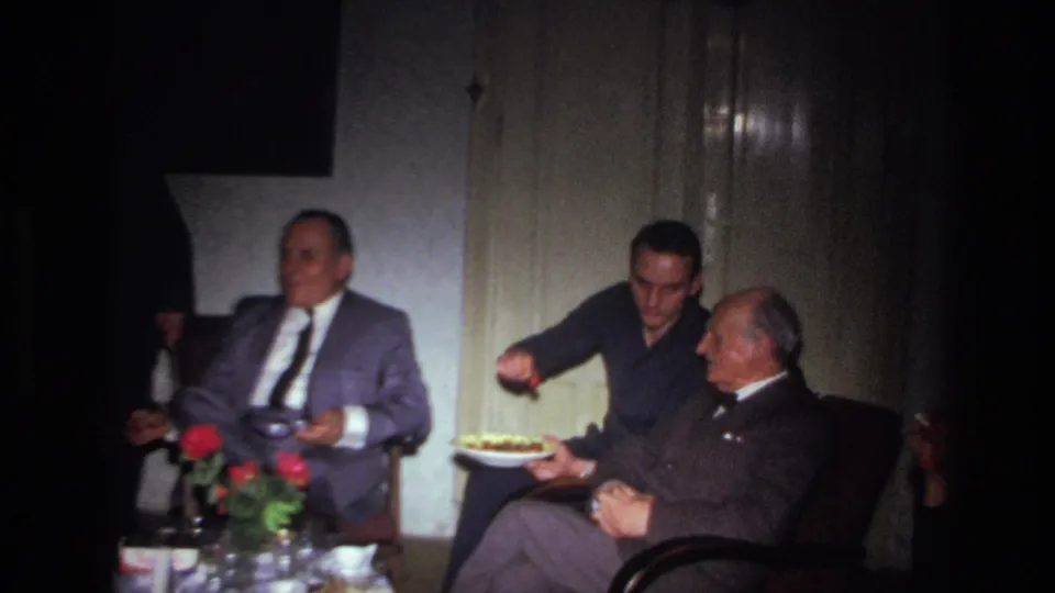 1967 France home-movie frame of adult men seated indoors, with a plate of food held between a younger man and an older man.