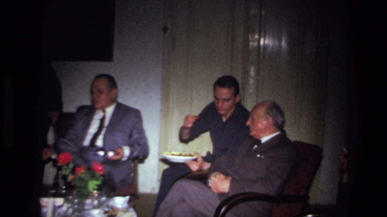 In 1967 France, three men sit in a dim private room as one holds a plate of food near a table with flowers and glassware.