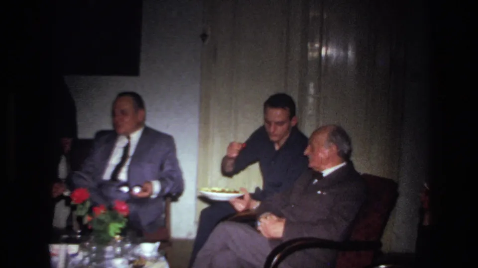 In 1967 France, three men sit in a dim private room as one holds a plate of food near a table with flowers and glassware.