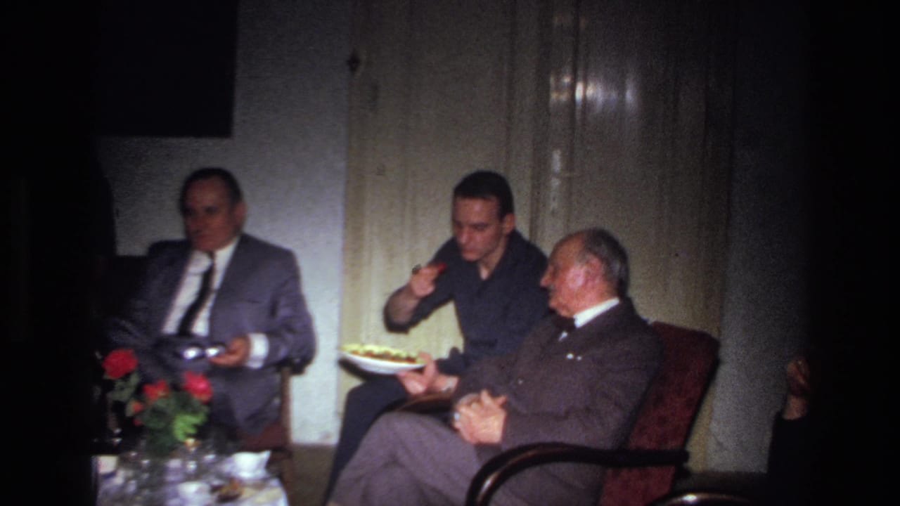 A man in a dark shirt serves food while two suited men sit beside him.