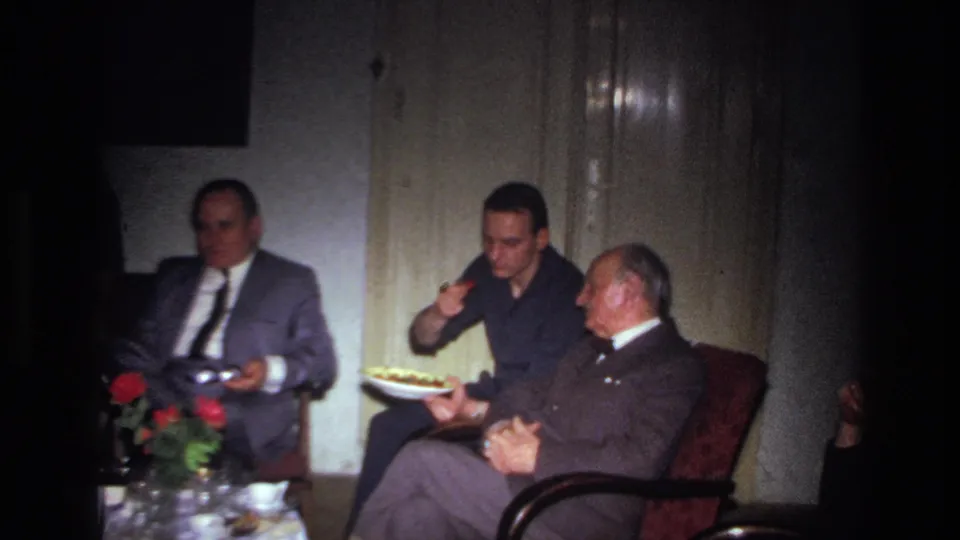 Three men in a private French interior, with the central man raising food from a plate while the older man watches.