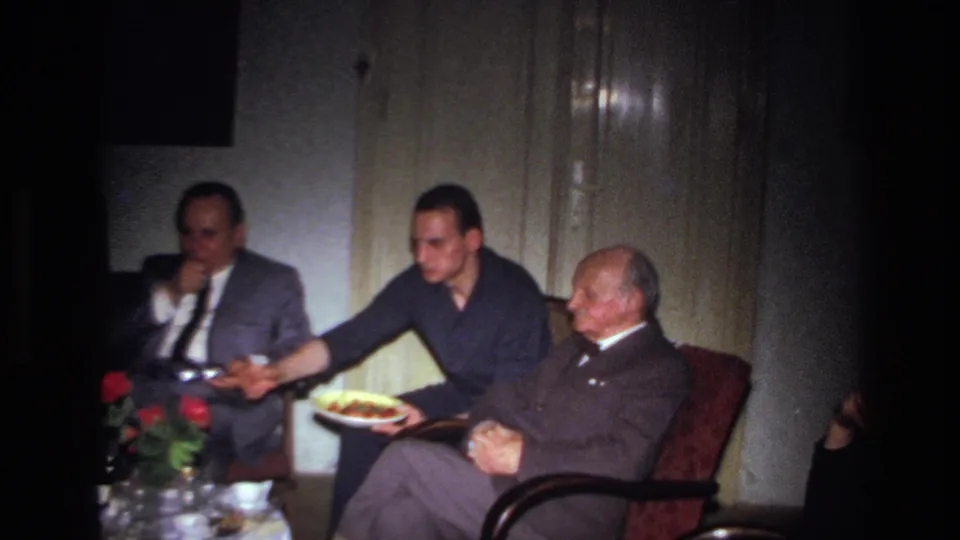 In the 1967 home-movie scene, the central man extends his arm toward a suited man while holding a plate in a dim room.