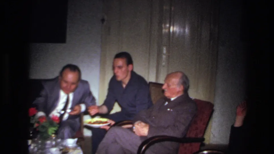 Three adult men sit in a dim French interior, one holding a plate of food as another leans forward near flowers and glassware.