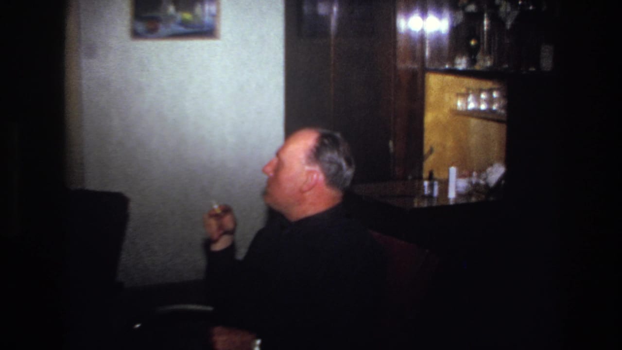 1967 France home-movie frame showing a man in a dark sweater smoking in profile beside a wooden cabinet and wall painting.
