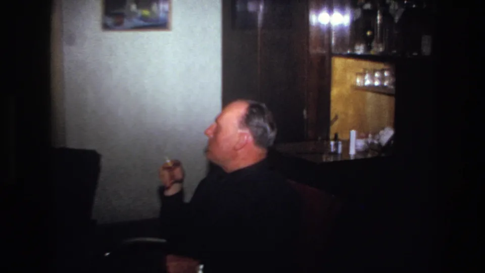 1967 France home-movie frame showing a man in a dark sweater smoking in profile beside a wooden cabinet and wall painting.