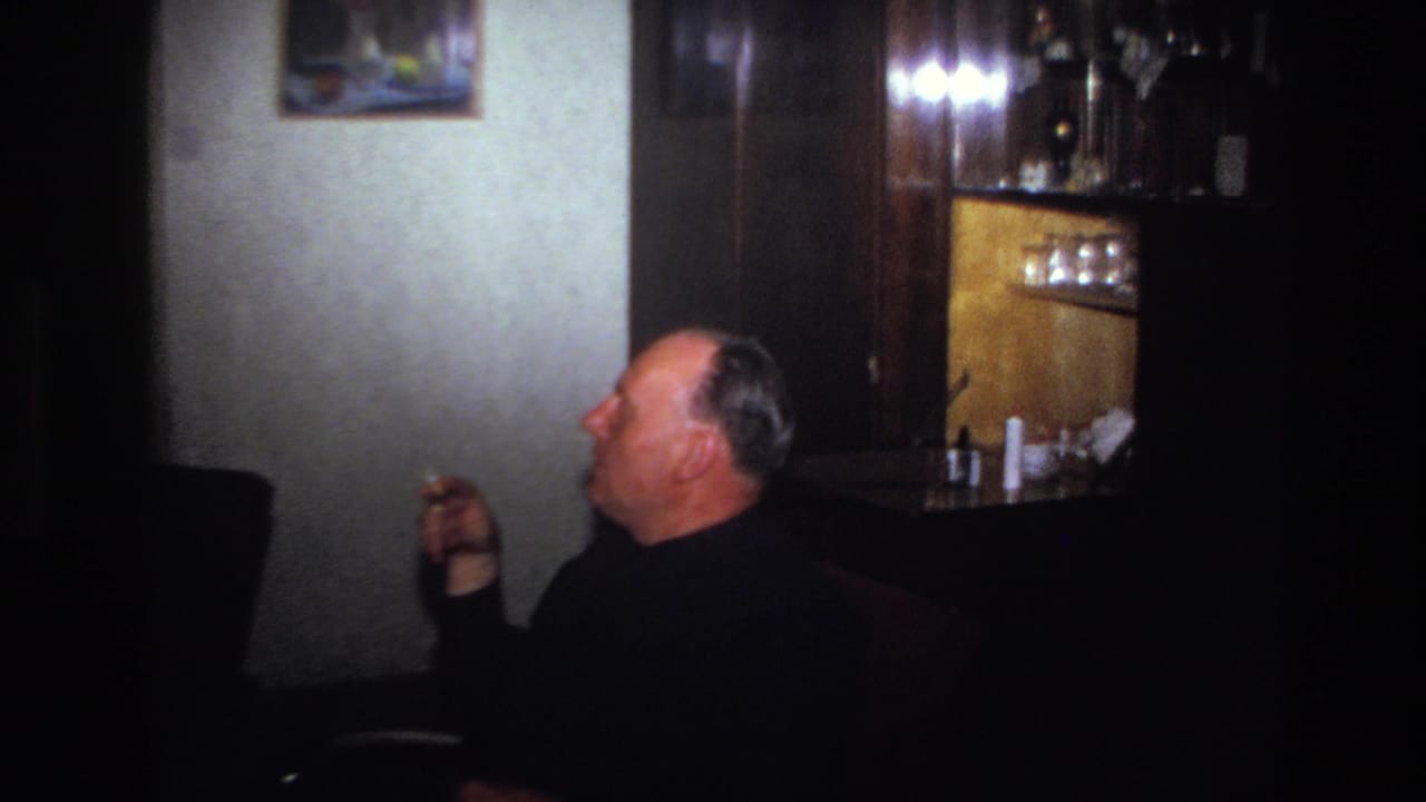 Man seated in profile with a cigarette near a cabinet and glassware.