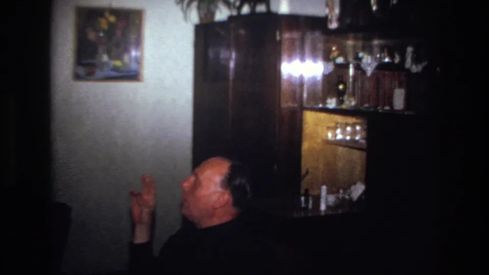 1967 France home-movie frame showing the man gesturing with a cigarette in a dim room with wood furniture and a painting.