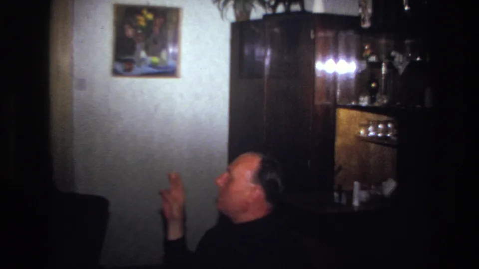 1967 France frame of a seated man turning and gesturing with a cigarette near a wooden cabinet in a private interior.
