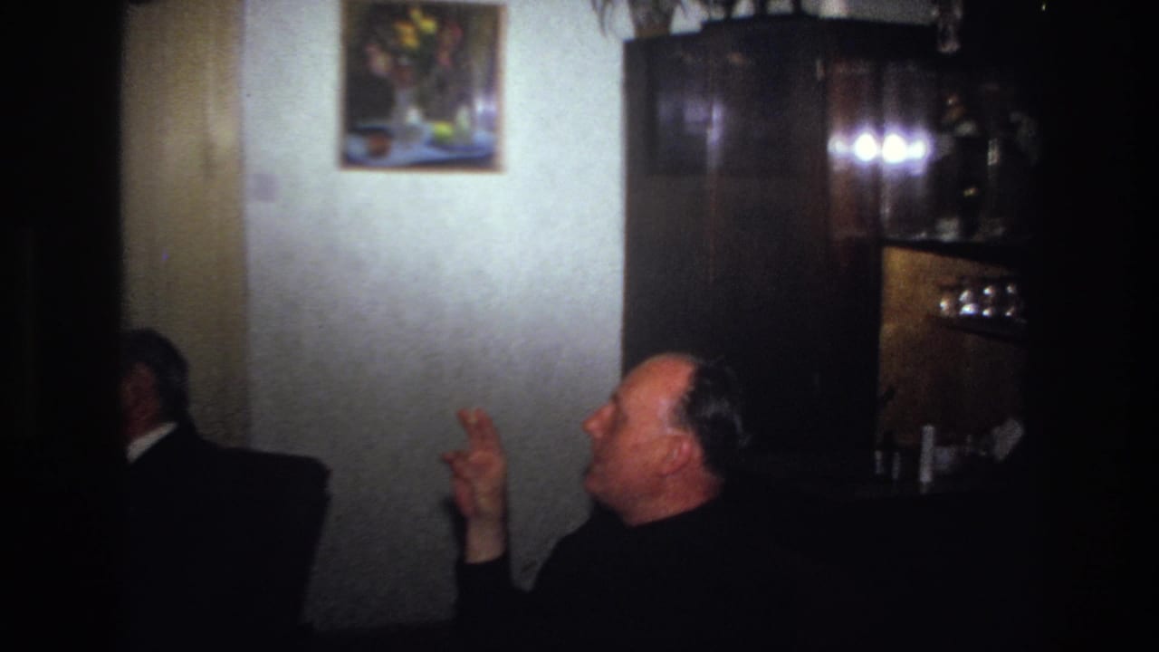 France 1967 home-movie frame showing well-dressed men seated in a dim private living room, with one man gesturing as he smokes.