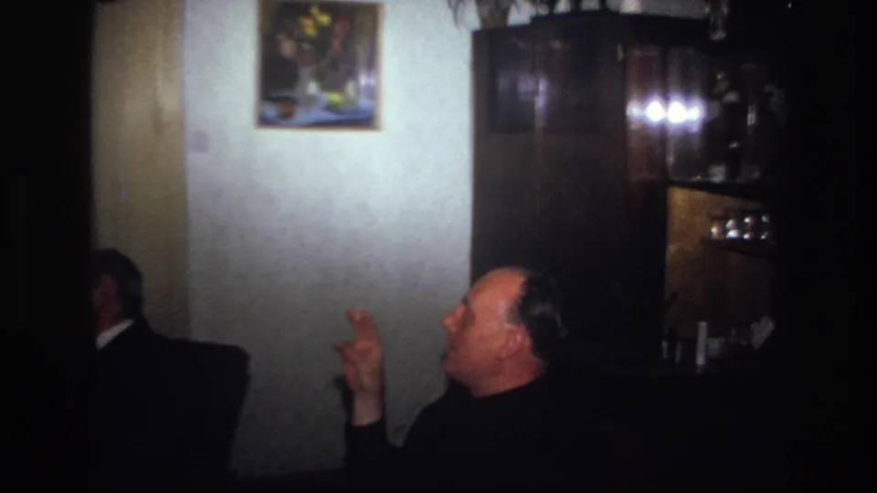 France 1967 home-movie frame showing well-dressed men seated in a dim private living room, with one man gesturing as he smokes.