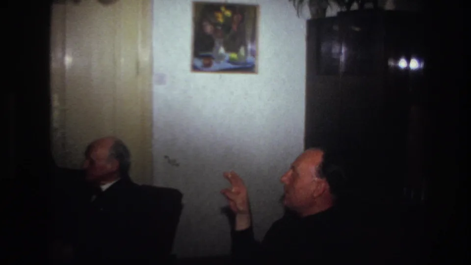 France 1967 frame with older men in dark clothing seated in profile near a wooden cabinet and framed flower painting.
