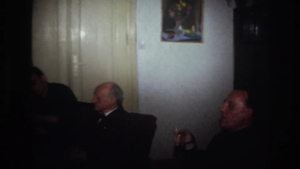 France 1967 home-movie frame showing three men seated in a low-lit living room, with a smoking gesture in the foreground.