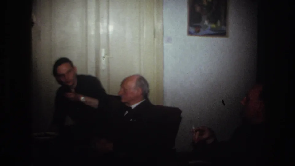 France 1967 frame showing a man leaning into a living-room conversation with seated older men and a smoker at the edge.