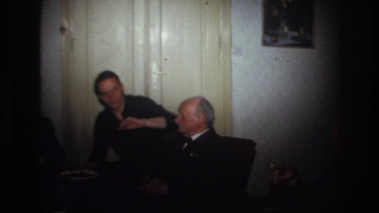 1967 France home-movie frame of adult men seated indoors as a younger man gestures toward an older man beside a pale door.