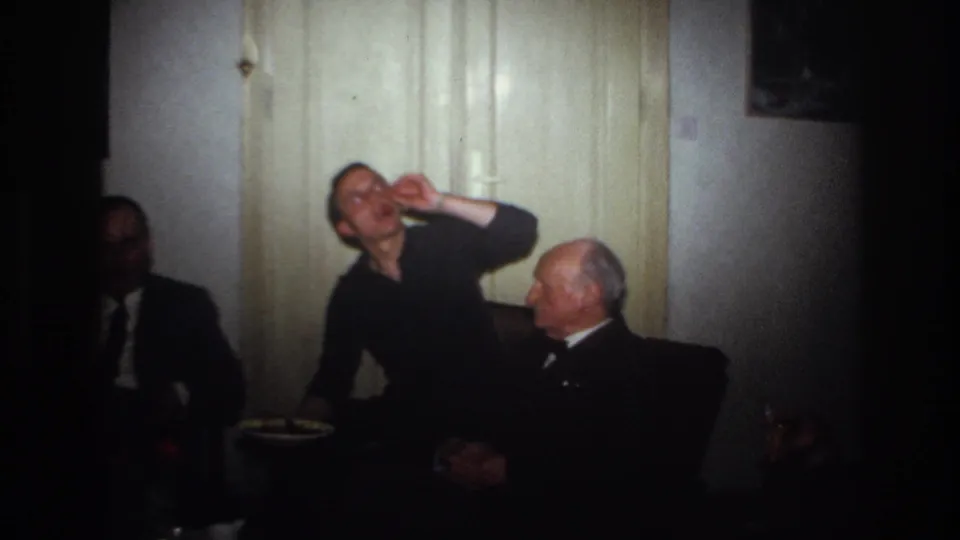 1967 France home-movie frame showing a seated man gesturing near his mouth as an older suited man looks on indoors.