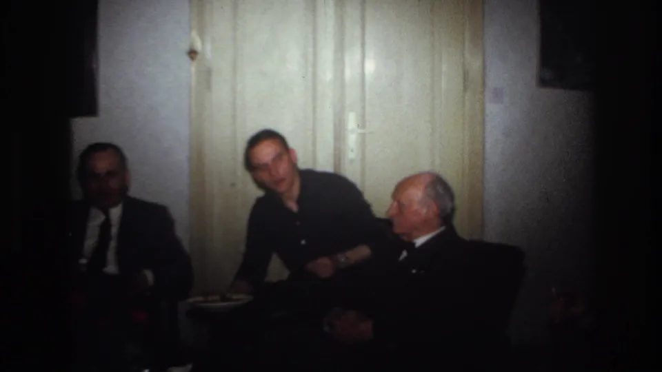 1967 France home-movie frame of three adult men seated in a dim private room while the central man leans into conversation.