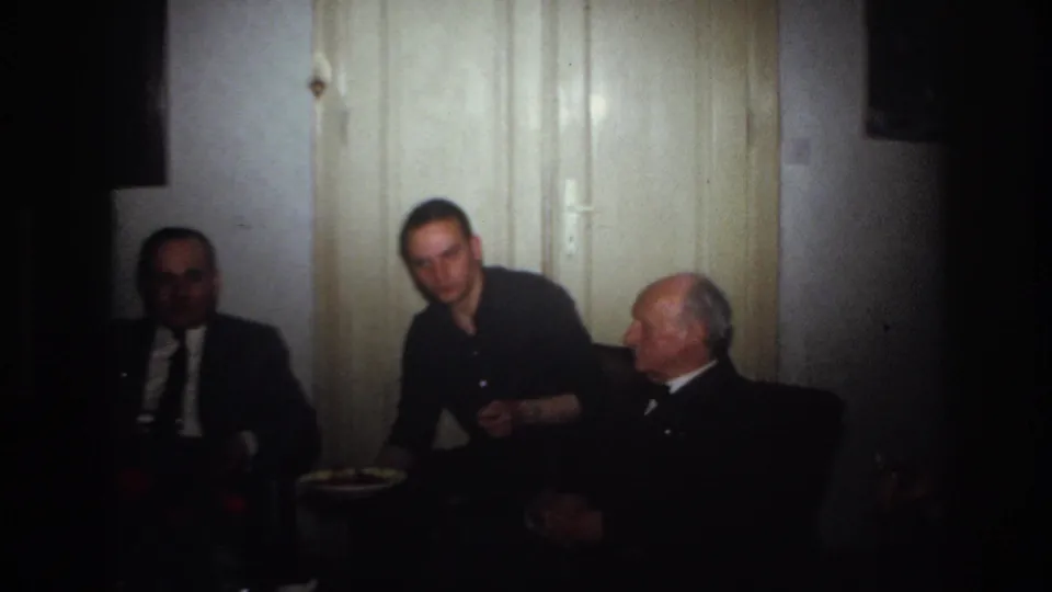 1967 France home-movie frame with a younger man facing forward between two seated suited men in a private interior.