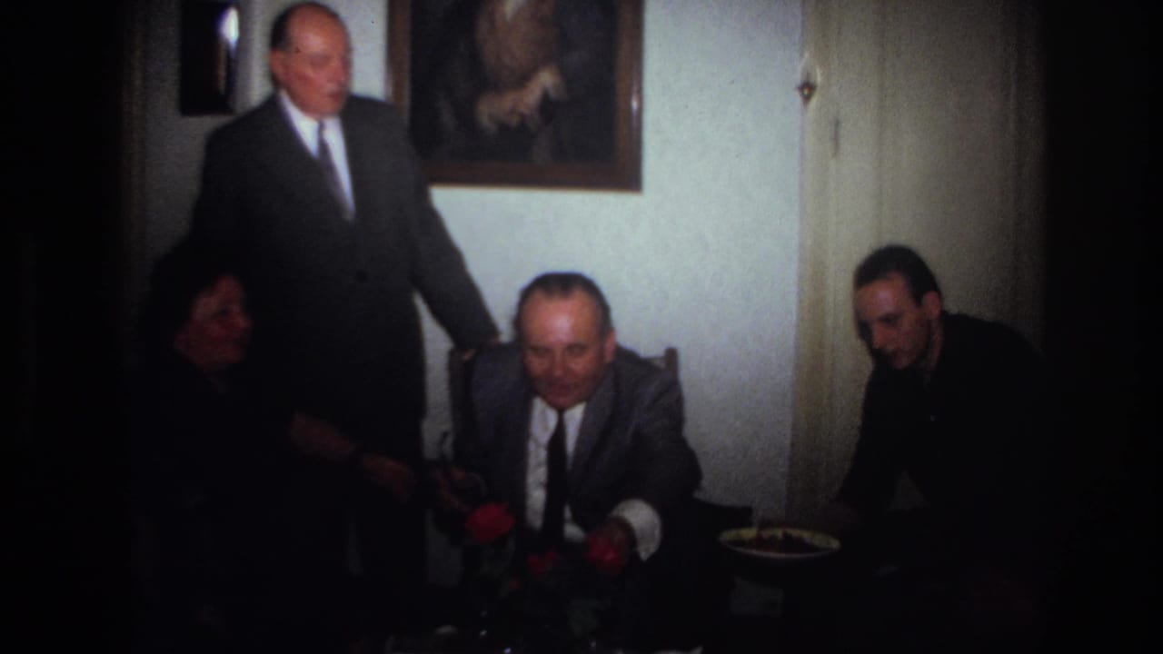 1967 France home-movie frame showing seated relatives in a dim private room, with a suited man standing behind them.