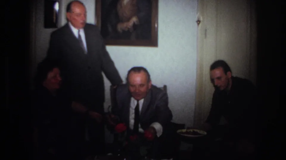 1967 France home-movie frame showing seated relatives in a dim private room, with a suited man standing behind them.