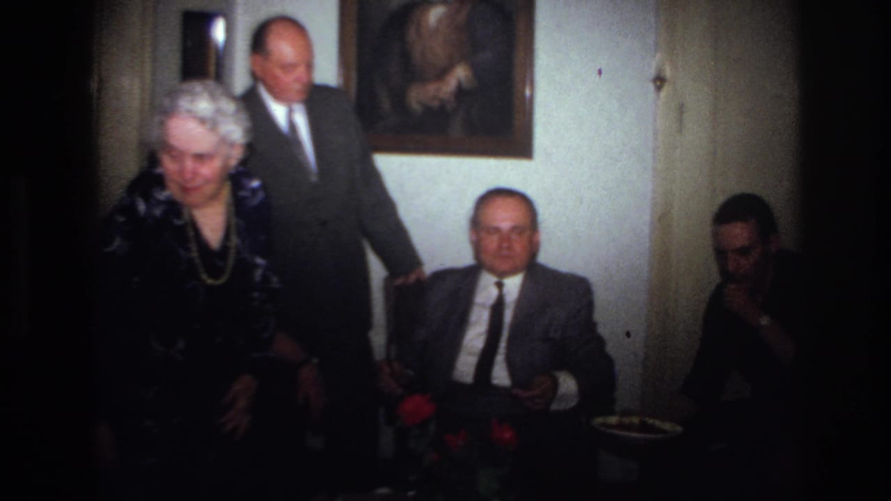 1967 France frame with an elderly woman in a dark patterned dress and necklace moving past seated family members.