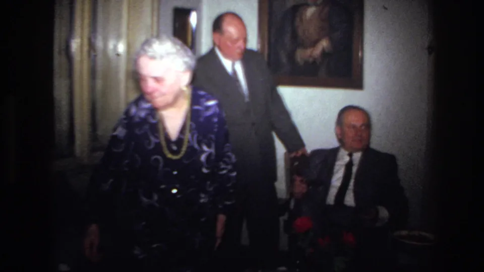 1967 France home-movie frame showing an elderly woman crossing the foreground of a family interior with seated relatives.