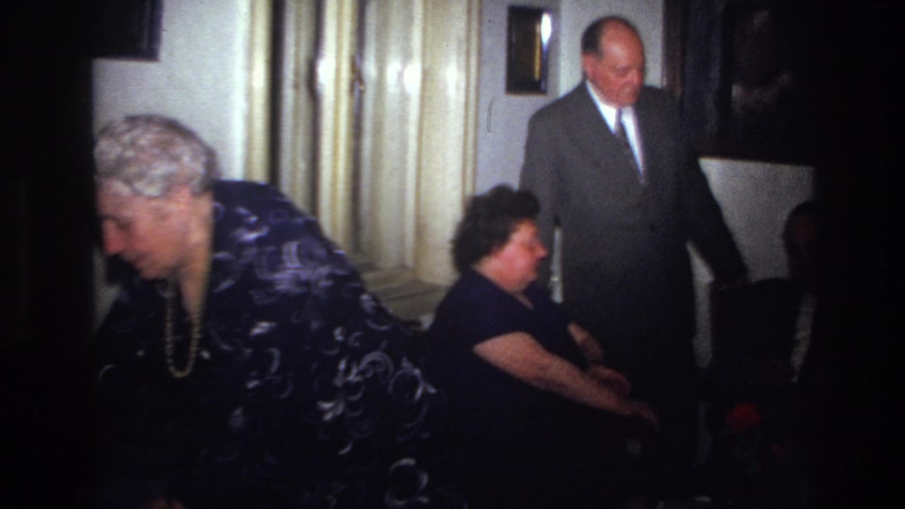 In a 1967 France home-movie frame, an older woman in a patterned dress leans forward as two suited men and another seated guest occupy a small interior.