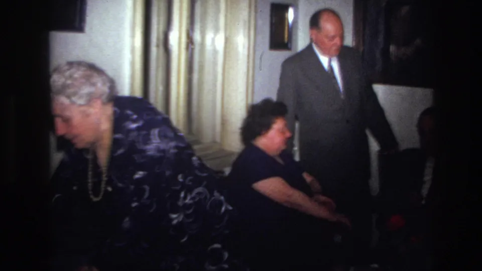 In a 1967 France home-movie frame, an older woman in a patterned dress leans forward as two suited men and another seated guest occupy a small interior.