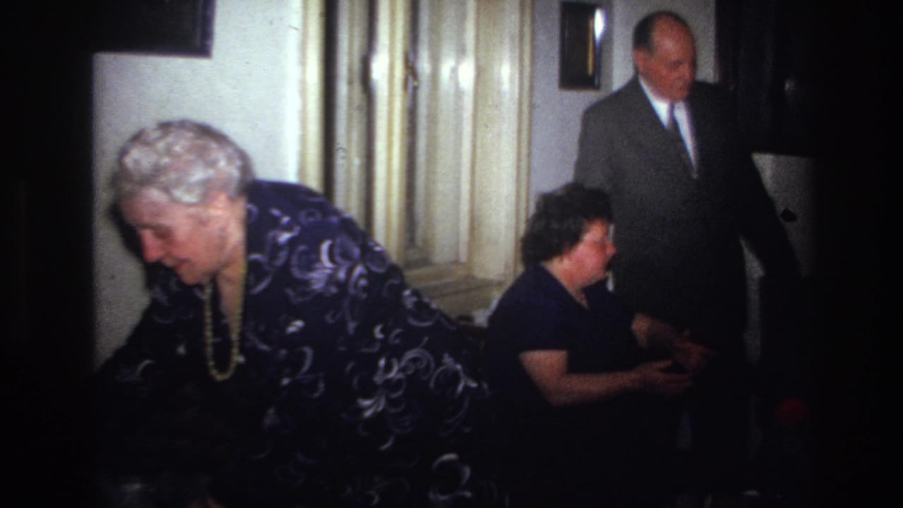 The home-movie frame shows an older woman bending forward in a private French interior while a suited man stands and a seated guest looks on.