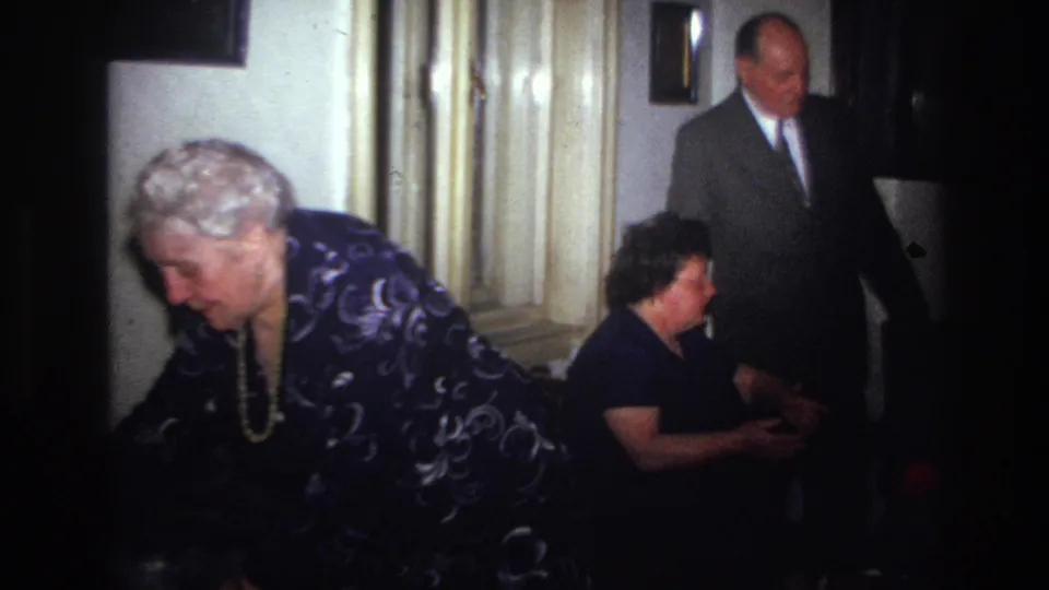 The home-movie frame shows an older woman bending forward in a private French interior while a suited man stands and a seated guest looks on.