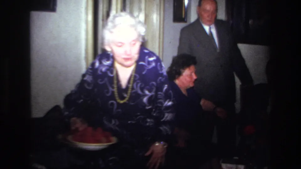 In this 1967 France interior frame, an older woman carries a bowl of red food past seated guests, with a suited man partly visible behind her.