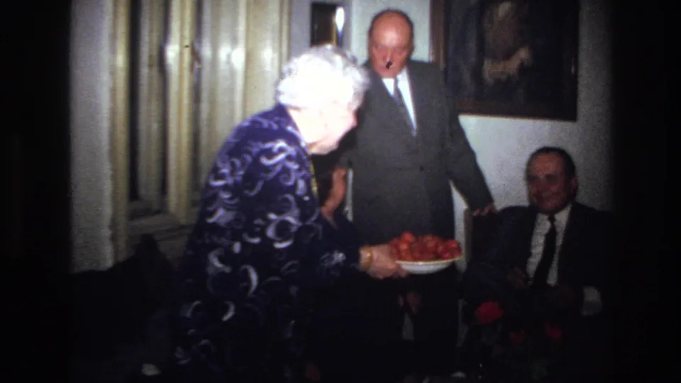 A 1967 France home-movie frame shows an older woman offering a bowl of food to a seated suited man while another suited man stands nearby.