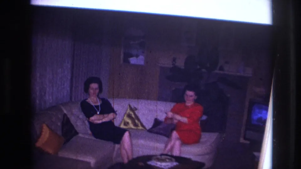 Two adult women sit on a white couch in a 1967 Ottawa home-movie frame, with patterned pillows, wood paneling, and a television nearby.
