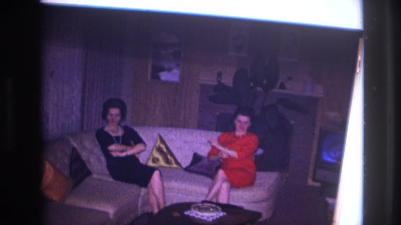 Two women sit on a light sofa in a wood-paneled living room with pillows and a small television nearby.